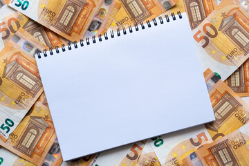 A blank page of a notebook lies on a background of money. Orange banknotes in denomination of 50 euros. Flat lay, copy space, top view.