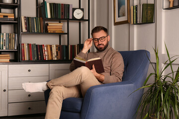 Young man reading book in armchair indoors. Home library