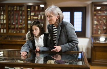 Friendly aged female tutor helping preteen girl exploring antique showpieces in local history museum..