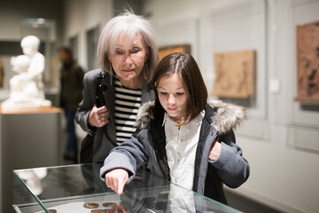 Friendly aged female tutor helping preteen girl exploring antique showpieces in local history museum..