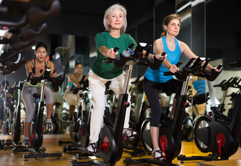 Two mature woman training on fitness bikes during cycling class in modern gym