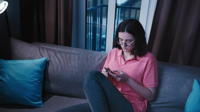 teen alone girl sits in the sofa near window in the evening and uses the phone, an evening alone, chat with friends, passing cars