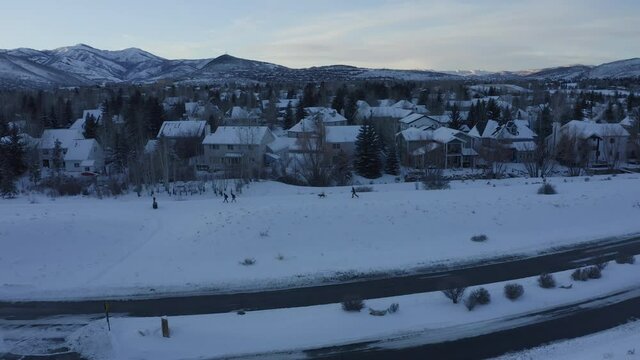 Drone view flying over thick layers of snow by the side of the road around Park City in Utah during winter. People walking back home at sunset with hills in the background.