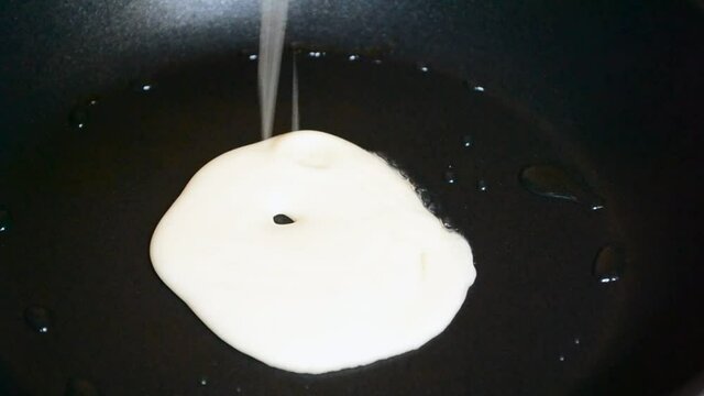 Liquid pancake mix gets poured into a black greasy frying pan. Person swinging the pan around in order to make a flatter pancake.