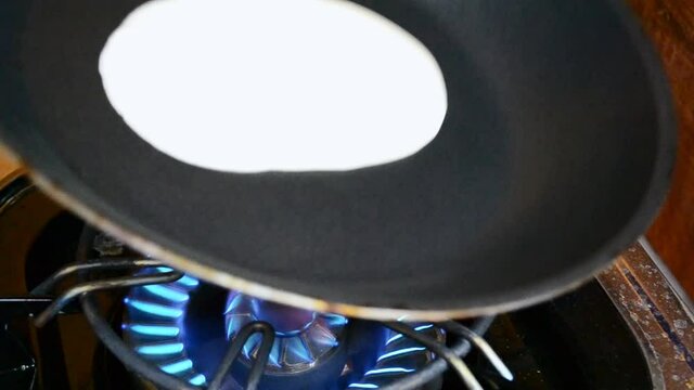 Liquid pancake mix is being poured into a black frying pan. Person swinging the pan around in order to make a flatter pancake.