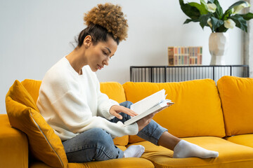 Fototapeta na wymiar Attractive black curly-haired woman reading her favorite historical novel sitting on a cozy yellow sofa