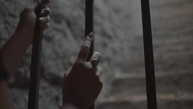 The manicured hands of a young woman hanging on the bars of a cell in the basement of an old building