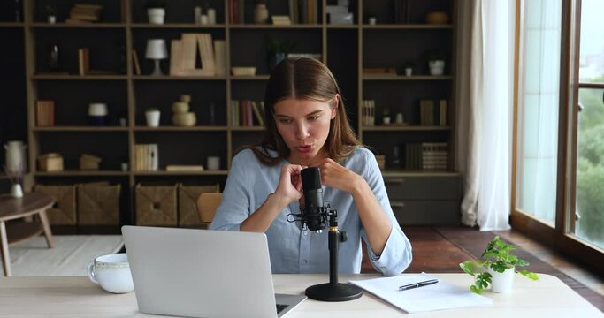 Attractive young woman sit at desk in cozy room makes speech into mike on stand, look at laptop take part in online stream topics, radio host work, modern tech, entertainment and communication concept