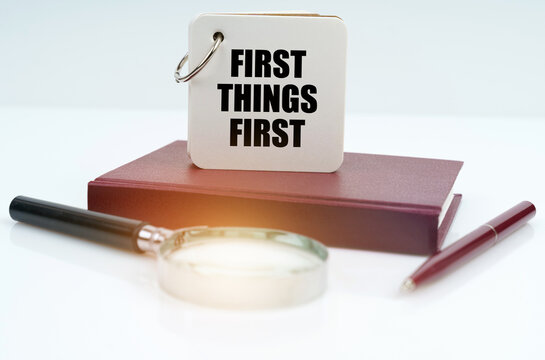 On a white surface lies a red notebook, a pen, a magnifying glass and a notebook with the inscription First Things First