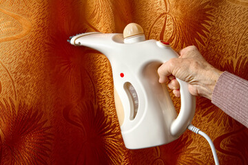 An electric steamer of white color in the hand of an elderly woman. - obrazy, fototapety, plakaty