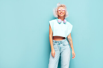 Young beautiful smiling female in trendy summer clothes. Sexy carefree woman posing near blue wall in studio. Positive blond model having fun and going crazy. Listening music in wireless headphones