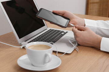 Woman connecting charging cable to smartphone near laptop at table indoors, closeup