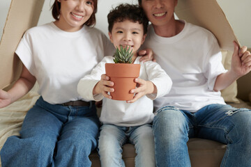 Smiling little kid with succulent plat and his happy parents sitting under cardboard roof, mortgage concept