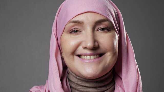 Young Muslim woman in light pink-colored hijab smiles widely posing for camera with confidence in studio room at professional illumination closeup.