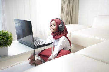 Asian moslem little girl wearing red hijab doing something on her laptop with headphone, for school activity.