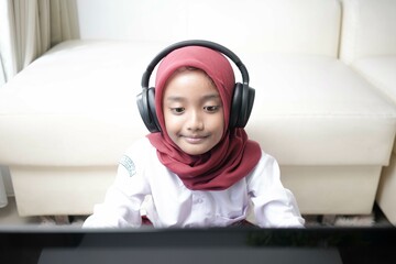 Asian moslem little girl wearing red hijab doing something on her laptop with headphone, for school activity.