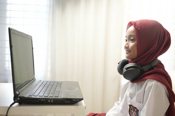 Asian moslem little girl wearing red hijab doing something on her laptop with headphone, for school activity.