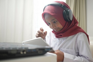 a cute little girl wearing red hijab doing something on her tablet computer and laptop. and wearing headphone