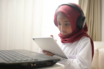 a cute little girl wearing red hijab doing something on her tablet computer and laptop. and wearing headphone