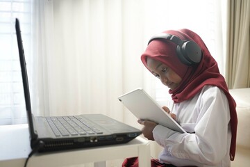 a cute little girl wearing red hijab doing something on her tablet computer and laptop. and wearing headphone