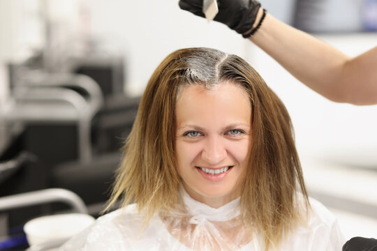 Happy client sit at hairdresser, master apply dye colour on hair with brush equipment