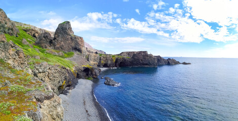 Fototapeta premium Scenic view of rocky coast with green grass and blue ocean; Person standing on the rock in a distance with blue ocean, black sand beach and green coastline with rocks and cliffs and blue cloudy sky