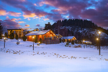 Winter landscape. Cottage on background of hill and forest. Wooden houses in countryside. New year in cottages. Country hotels Christmas evening. Renting house for large group. Winter village