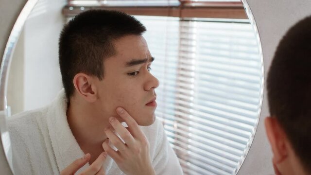 Unhappy Asian Man Squeezing Pimples Looking At Mirror In Bathroom