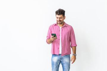 Fototapeta premium Confident young handsome man in jeans shirt holding smart phone and looking at it with smile while standing against white background