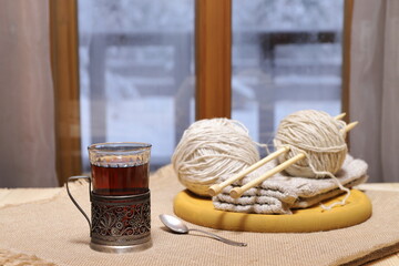 A glass of tea, woolen threads and knits on the table in front of the window on the background of...