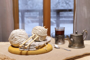 Fototapeta na wymiar A woolen threads and knits, glass of tea on the table in front of the window on the background of winter landscape.