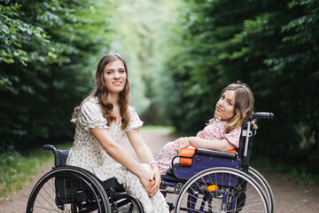 Portrait of young caucasian ladies in wheelchairs sitting in front of each other at summer park and...