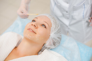Obraz na płótnie Canvas Hands of cosmetology specialist applying facial mask using stick, making skin hydrated and face glowing. Attractive woman relaxing smiling and enjoying spa procedures