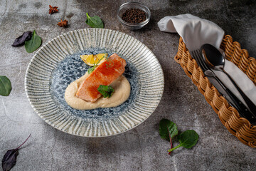 Serving a dish from a restaurant menu. Baked salmon steak with lemon, salad and dill and parsley greens on a plate against a gray stone table