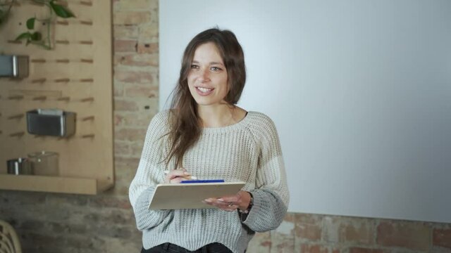 Female student presentation in classroom. Dark hair talking smiling and wearing winter clothes. Standing with the notebook in the modern classroom near the whiteboard and writing.