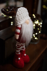 Ozdoba świąteczna, skrzat, krasnal, elf z białą brodą, czapką i porożem renifera. Ubrany w czerwono-białe ubranko w paski. W tle światełka choinkowe - obrazy, fototapety, plakaty