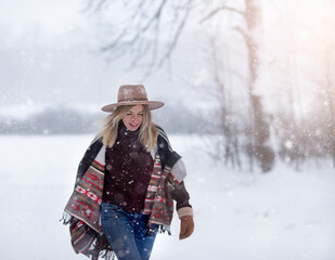 Fototapeta na wymiar Happy young woman walks through a snow-covered field. The forest is visible on the horizon. She is wearing a hat and a bright cape. Snow falls on her face, the wind blows her blonde hair.