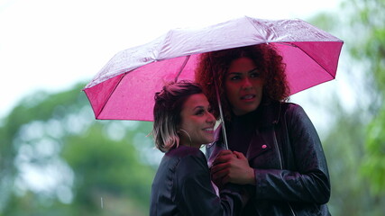 Two diverse friends under umbrella in the rain waiting for taxi