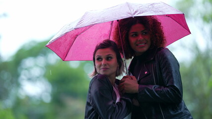 Two diverse friends under umbrella in the rain waiting for taxi