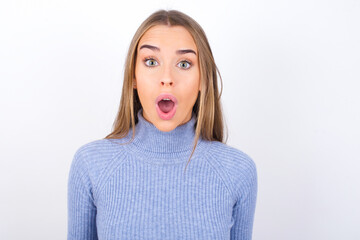 Young caucasian girl wearing blue turtleneck over white background having stunned and shocked look, with mouth open and jaw dropped exclaiming: Wow, I can't believe this. Surprise and shock