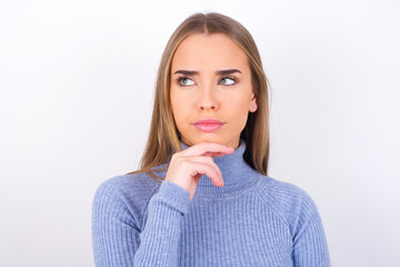 Shot of contemplative thoughtful Young caucasian girl wearing blue turtleneck over white background keeps hand under chin, looks thoughtfully upwards.