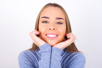 Dreamy Young caucasian girl wearing blue turtleneck over white background keeps hands pressed together under chin, looks with happy expression, has toothy smile.