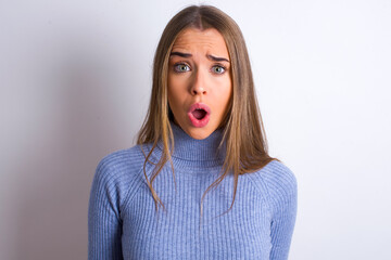Expressive facial expressions. Shocked stupefied Young caucasian girl wearing blue turtleneck over white background , keeps jaw dropped feels stunned from what he sees aside.