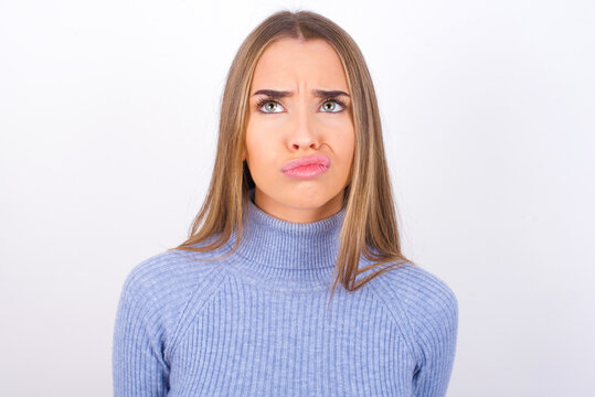 Young caucasian girl wearing blue turtleneck over white background crosses eyes, puts lips, makes grimace with awkward expression has fun alone, plays fool.