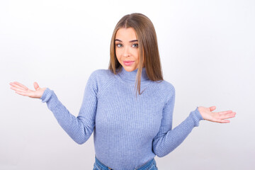 Puzzled and clueless Young caucasian girl wearing blue turtleneck over white background with arms out, shrugging shoulders, saying: who cares, so what, I don't know. Negative human emotions.