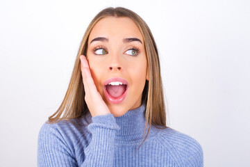 Young caucasian girl wearing blue turtleneck over white background excited looking to the side hand on face. Advertisement and amazement concept.