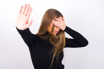 Young caucasian girl wearing black turtleneck over white background covers eyes with palm and doing...