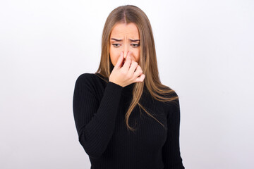 Young caucasian girl wearing black turtleneck over white background , holding his nose because of a bad smell. - obrazy, fototapety, plakaty