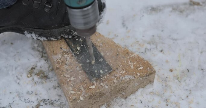 drilling an iron plank on a wooden board with a drill in the snow while holding the metal plate with your foot. Close-up