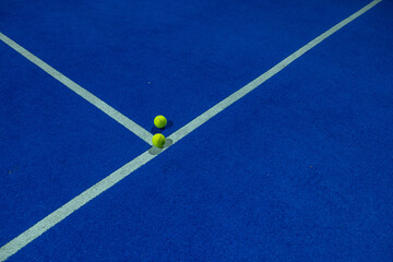 Two paddle tennis balls in the foreground on the line of a blue paddle tennis court at night.